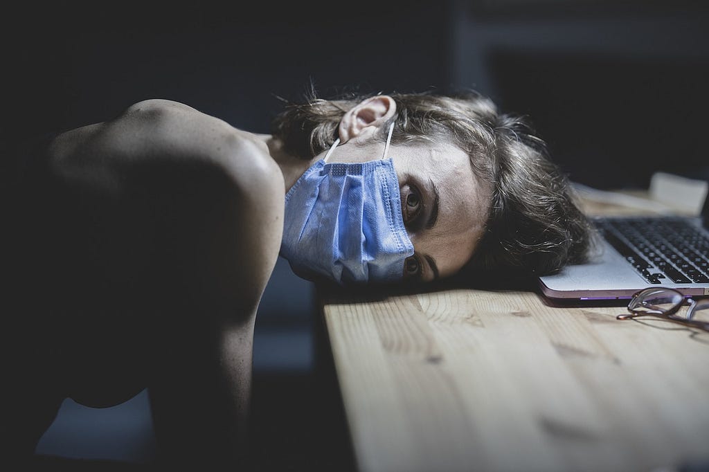 Registro fotográfico altamente conceptual: en un ambiente semi oscuro, una joven mujer usando máscara de protección contra el coronavirus y postrada sobre una mesa en la cual se ve un notebook y sus anteojos.