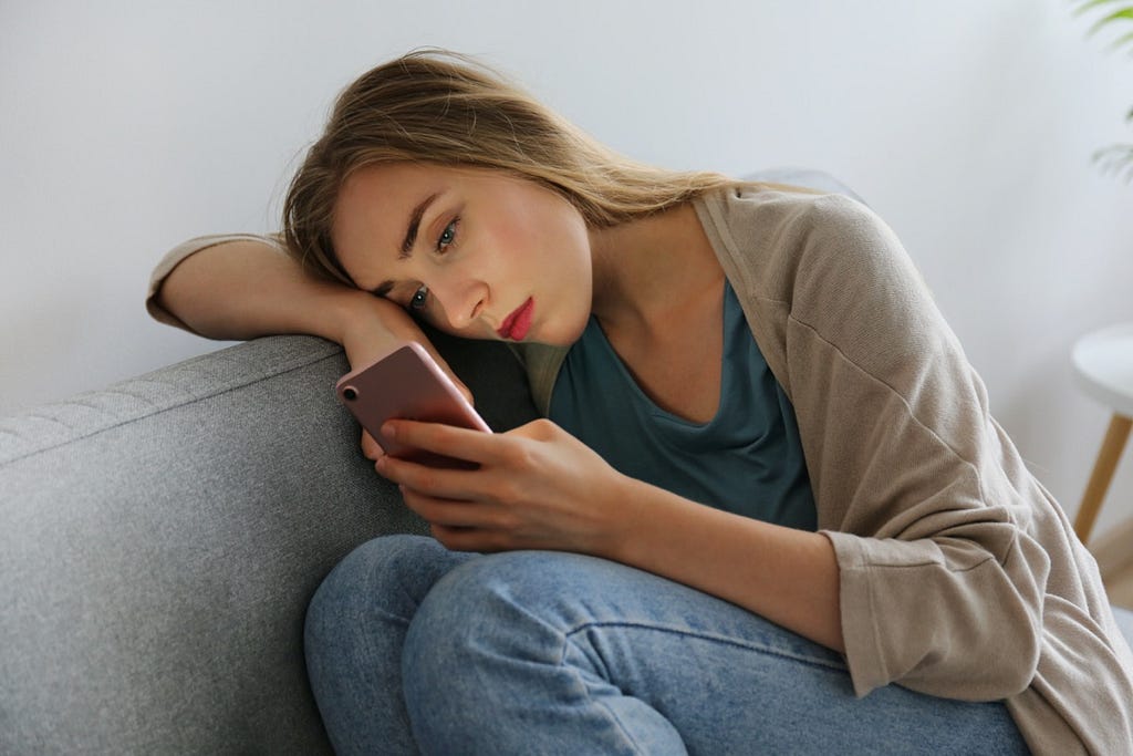 A photo of a girl staring at her phone, sitting alone on the couch and feeling sad.