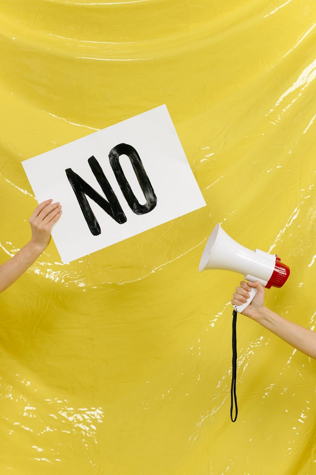 Hands of two individuals on a yellow background: one hand is holding a microphone and the other one is holding a banner written “no” on it in capital letters.