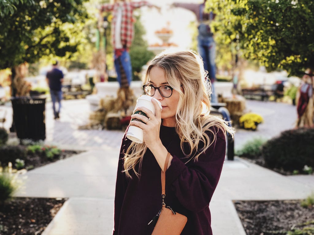 Blonde woman with eyeglasses sipping coffee while walking along a park
