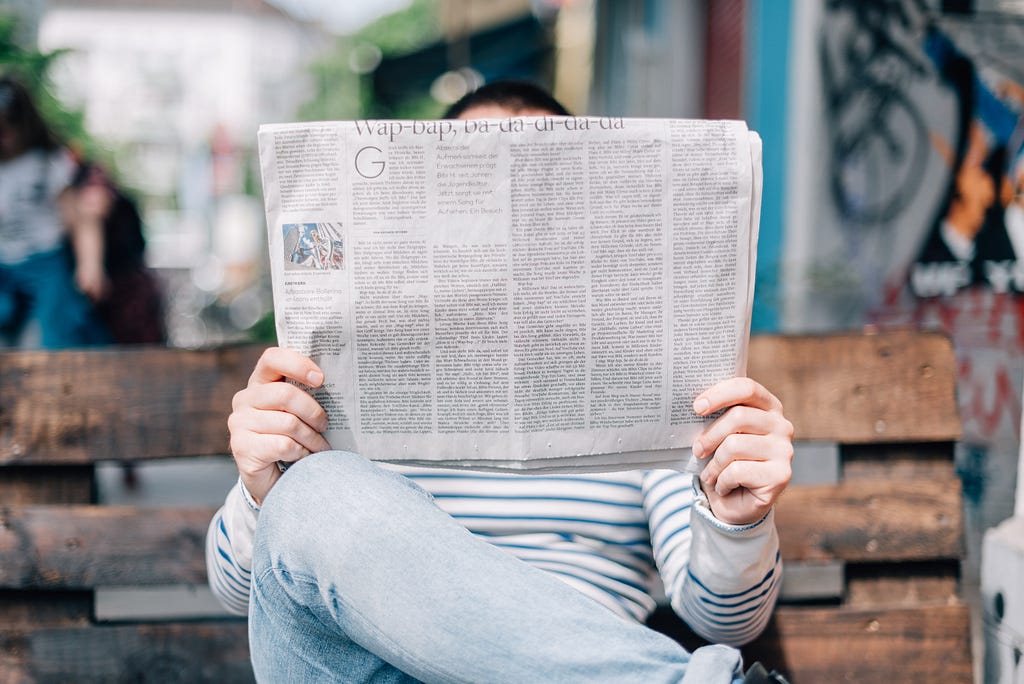 woman dressed in jeans — sitting on a wood bench — reading a newspaper