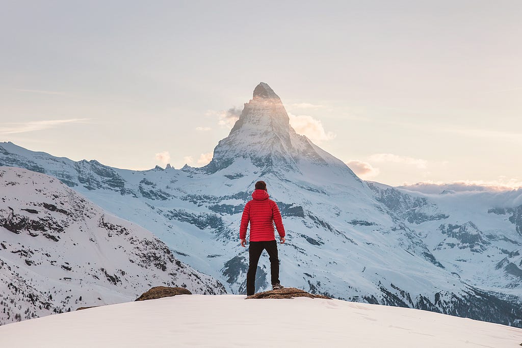 A man standing on a snowy mountain overlooking a higher mountain