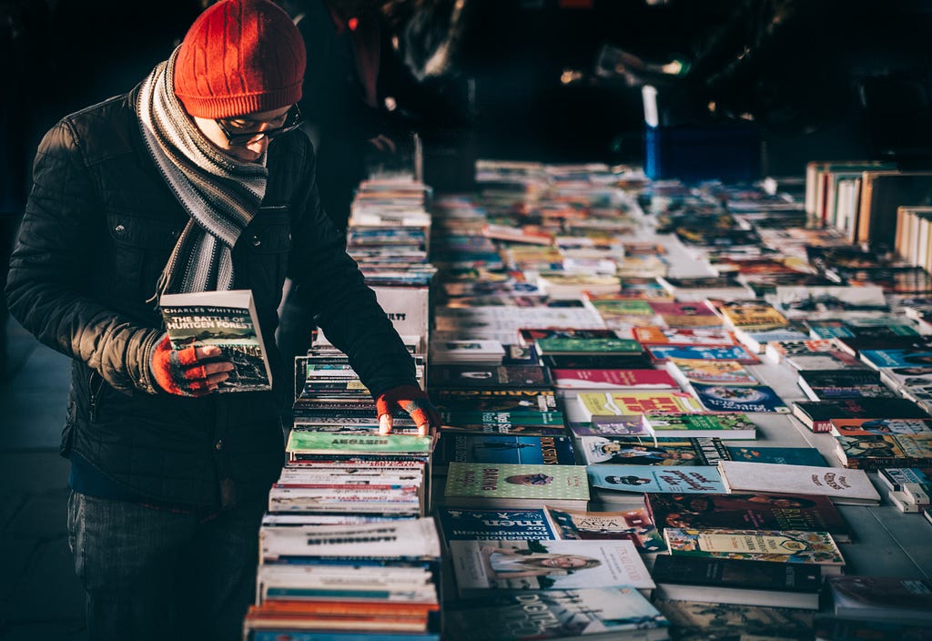 Person with cold weather attire is standing over a table full of books with one in hand.