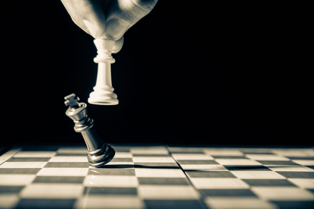 A dramatic close-up of a chessboard depicts the final moments of a match. A hand suspends a white Queen over a fallen black King, symbolizing victory and defeat. The stark contrast between light and dark, combined with the sharp focus on the pieces, emphasizes the intensity and strategic nature of the game. This powerful image evokes themes of competition, triumph, and the decisive role of leadership.