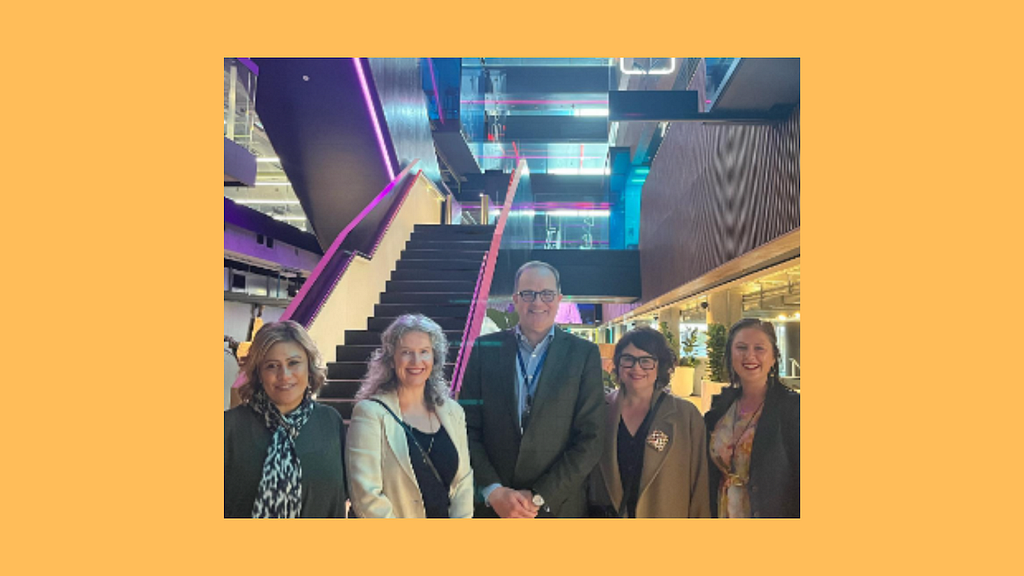 Image description: 4 members of Project Gender with TVNZ Chief Executive Simon Power standing in the middle look at the camera smiling. The background is the foyer of TVNZ including a large staircase lit by purple lighting.