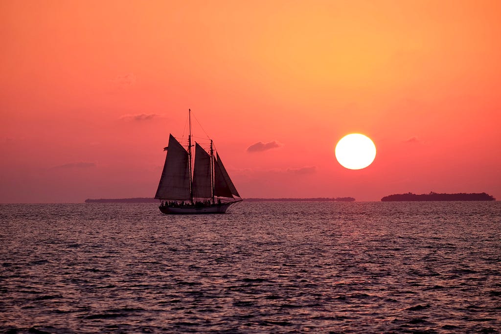 The silouette of a sailing ship on a large body of water. The sun sets behind it.