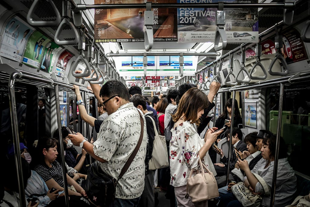 A crowded subway car with passengers standing and sitting, most of them looking at their smartphones, surrounded by advertisements and handrails.