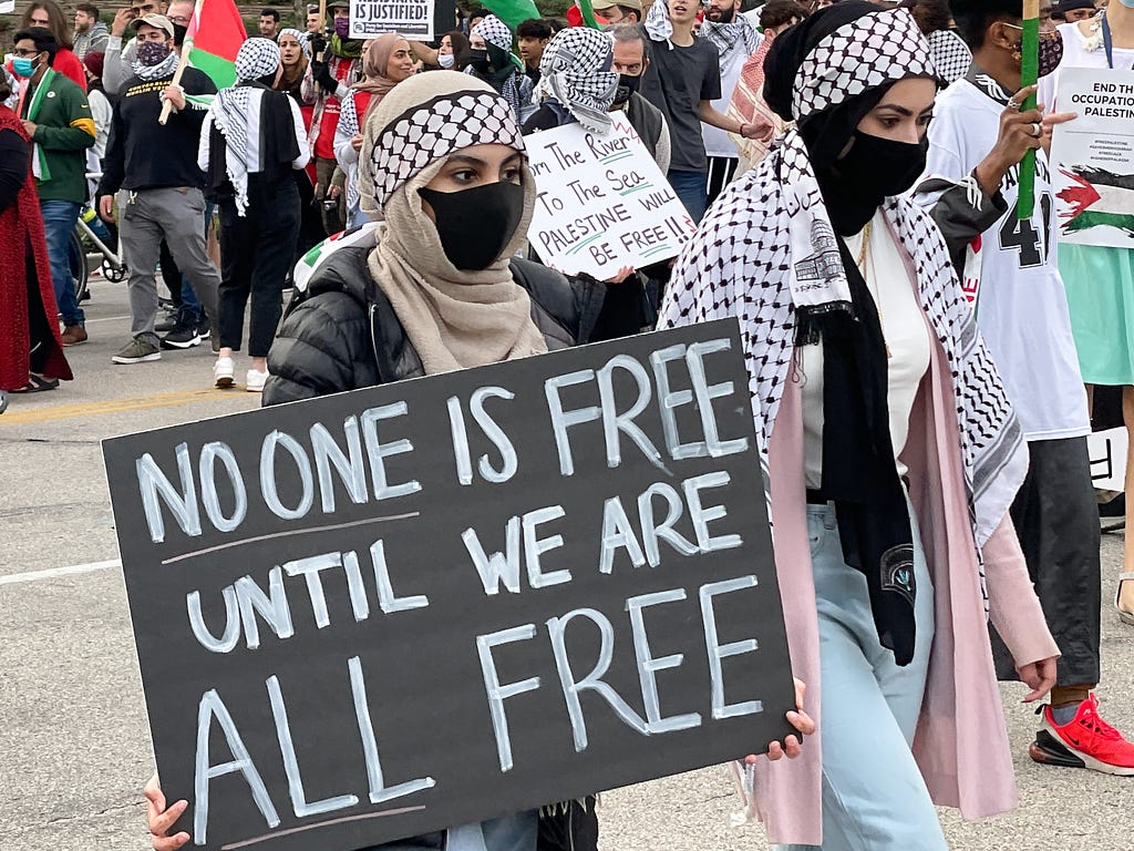 Detail from a march with various people wearing keffiyeh and holding signs about Palestine, focused on a person wearing hijab with a keffiyeh wrapped around it and holding a sign reading “no one is free until we are all free”
