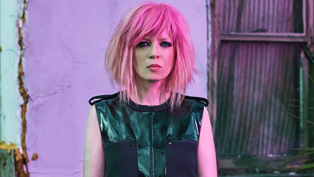 A photograph of Shirley Manson, lead singer of American band Garbage. She is standing in front of a backdrop of concrete and corrugated steel. She has pale skin, and neck-length pink hair. She is wearing a black sleeveless top with patches of different material and a front zipper. Her facial expression is neutral and unsmiling. She is looking directly at the camera.