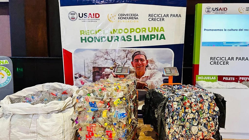 A man sits behind three large bags of crushed plastic bottles and cans. He sits in front of a large poster with logos that says Recycling for a Clean Honduras.