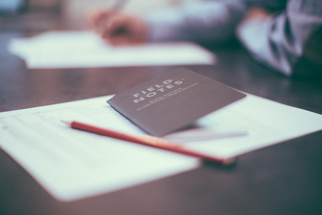 A notebook titled “field notes” and a pencil with paper atop a desk.