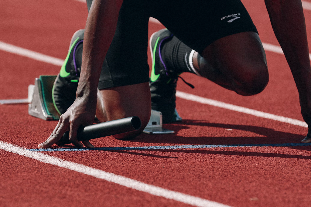 Pessoa negra em posição de largada, ela está agachada segurando um bastão em meio a uma pista de corrida.