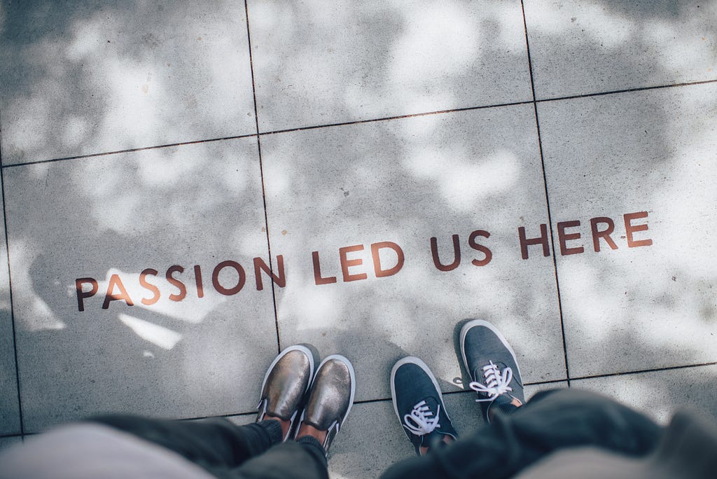 Two people only visible by their shoes at the bottom of the image with the phrase” Passion led us here” painted on the concrete in front of them