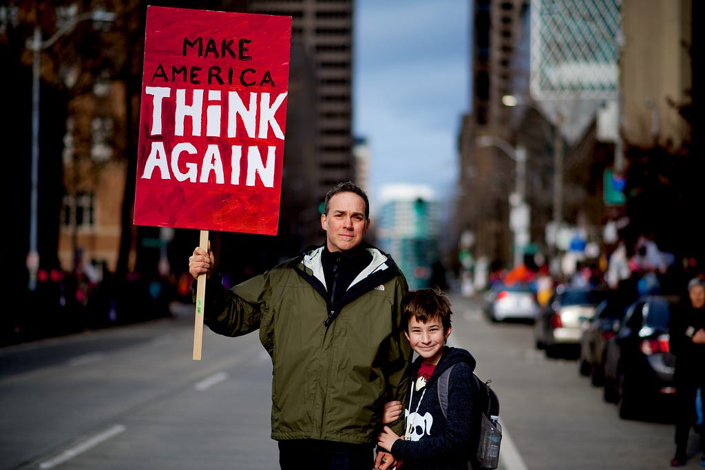 A father, holding a board printed with “Make America Think Again,” is standing with his son.