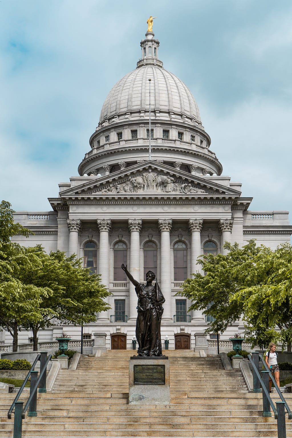 Light grey building that looks like a domed state capitol building, with pillars, trees, a grand statue on a pedestal in front, and a golden statue atop the dome.