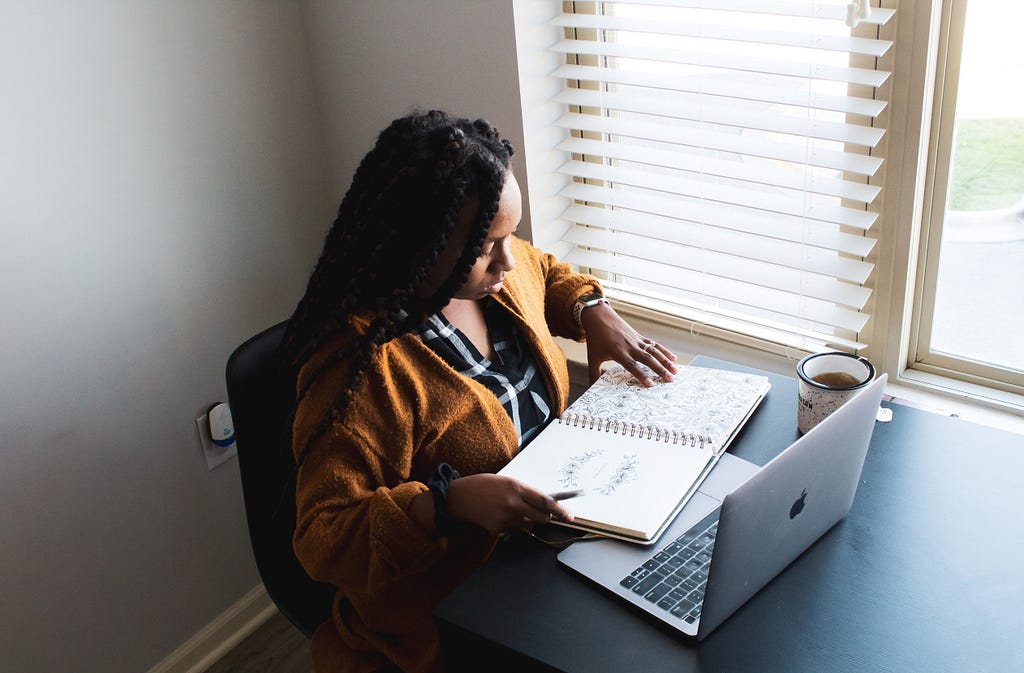 A black woman writing on a notepad at a desk with her laptop