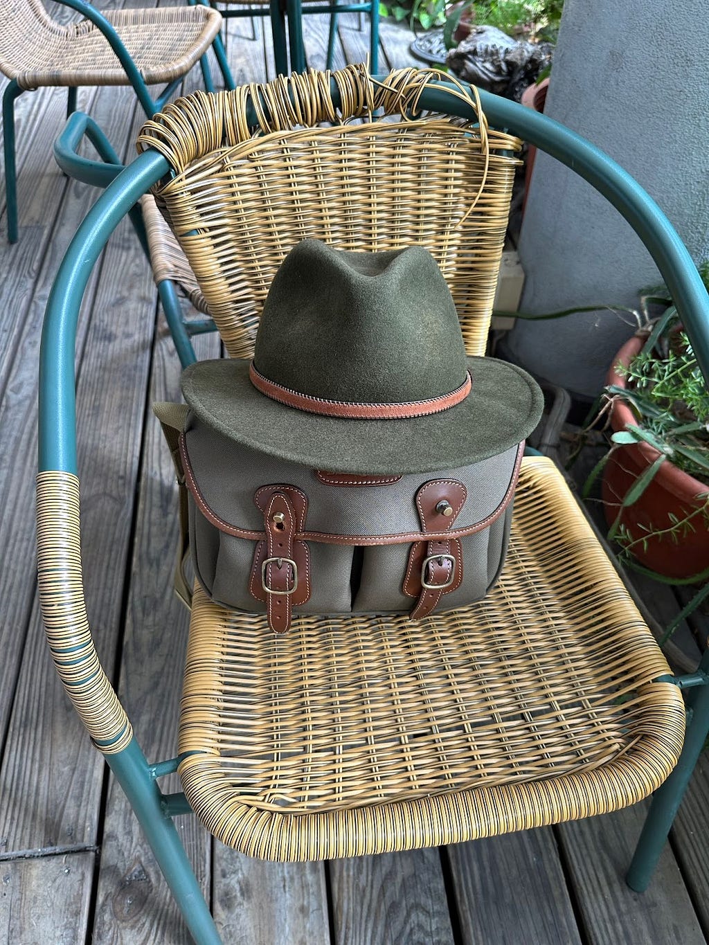 Wicker chair with small sage green satchel/bag with brown leather trim and straps resting on it. Green felt fedora on top of the bag.