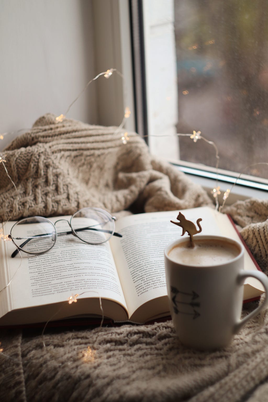 A cup of latte with cat spoon, an opened book, a glasses, a sweet brown cardigan, placed by the side of window. There’s also a cute small star wire-light over them