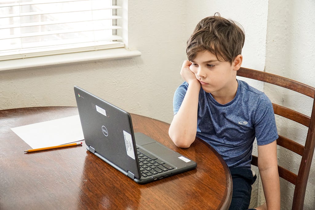 A young boy resting his hand on his face and his elbow on a wooden table, seated in front of a laptop.