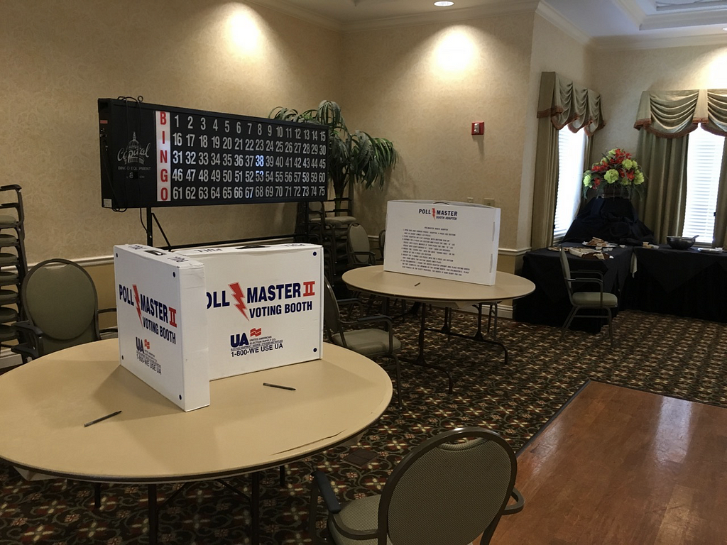 Large room with 2 round tables and boxes arranged to make privacy booths for voting — with a large Bingo board in background