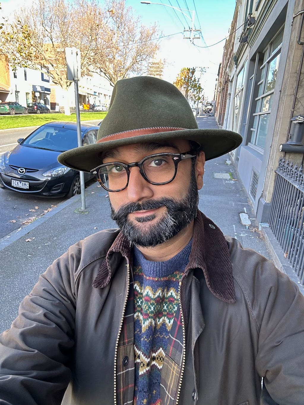 Selfie of brown man with beard, glasses, green felt fedora, wearing waxed cotton jacket. Standing in street with parked cars.