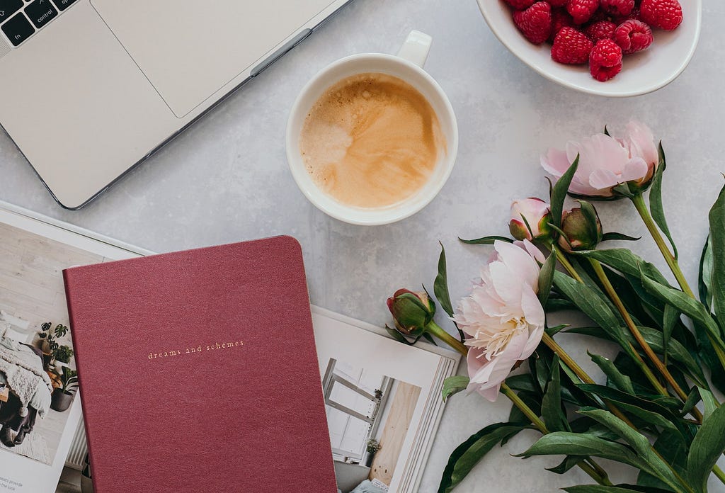 Desk with laptop, pink flowers, a notebook ,a cup of coffee and some ruspberries