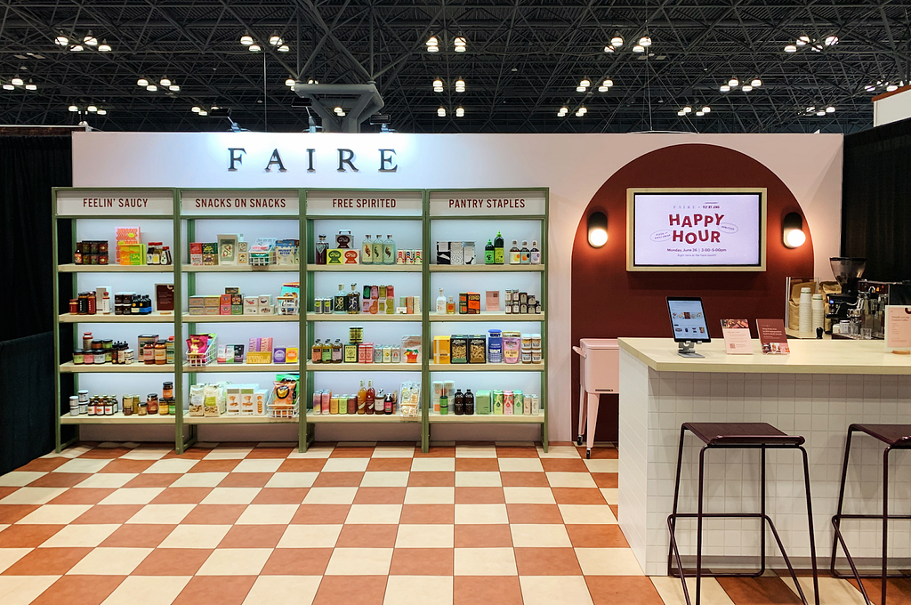 A photo from Faire’s booth at Summer Fancy Food. There is a checkerboard print floor, four shelves stocked with products that look like bodega shelves divided according to category (Feelin’ Saucy, Snacks on Snacks, Free Spirited, and Pantry Staples), a screen with the text ‘Happy Hour’, and a counter with a tablet and two chairs.