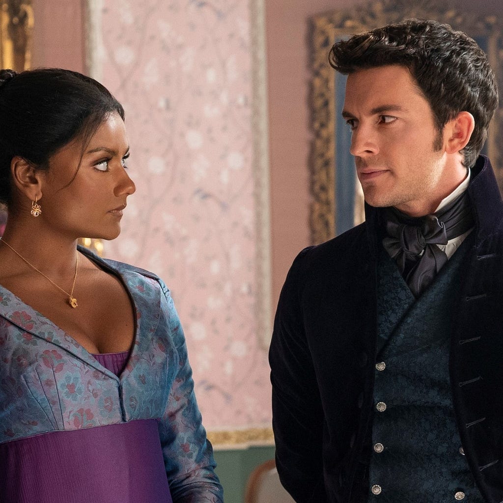 An Image shows Simone Ashley as Kate Sharma and Jonathan Bailey as Anthony Bridgerton in Bridgerton Season 2. The actors are in period clothing in the colours blue and violet. They look at each other.