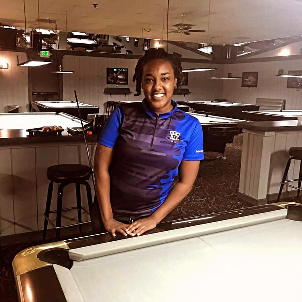 The author, Amani Ali, poses with her hands resting on the edge of a pool table with grey cloth in an empty pool room.