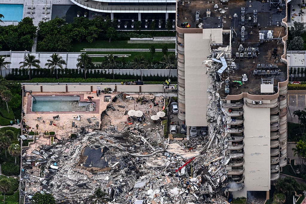 An aerial photo of the partially collapsed condominium. An entire face of the building is missing and debris is scattered below, crushing the surrounding area.