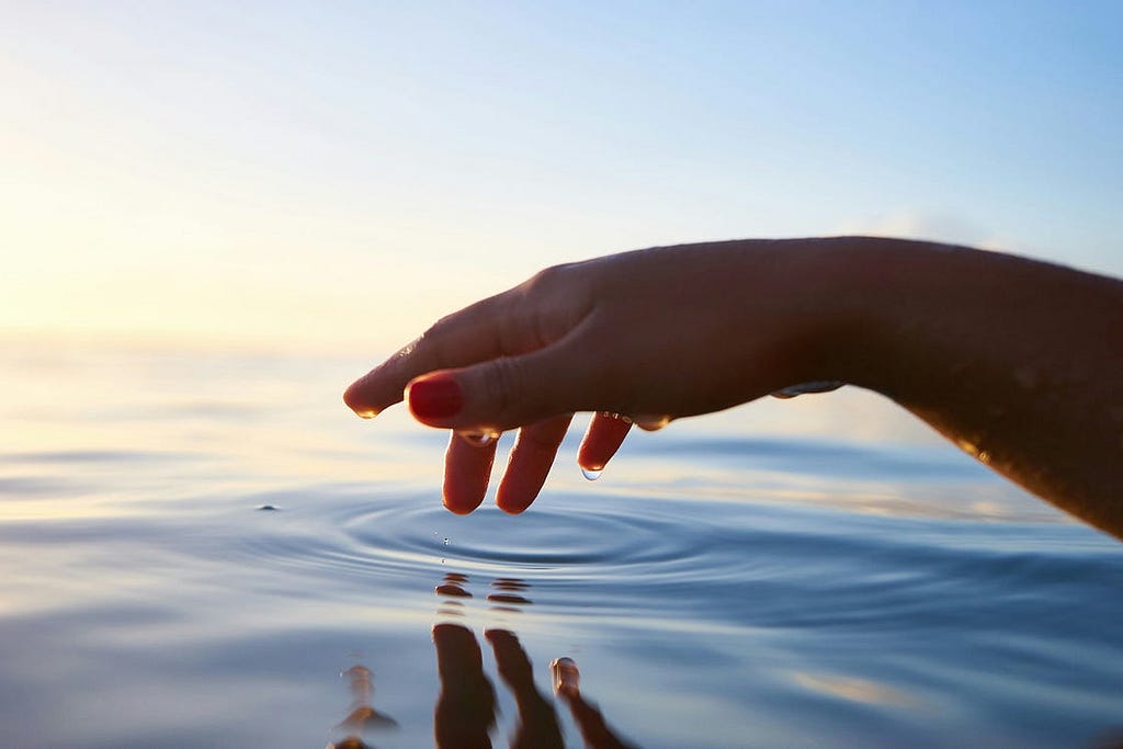 An open water scene. There is a hand coming from the right making a ripple in the water. There is a faded yellow/white light coming from the left in the background on a blue sky.