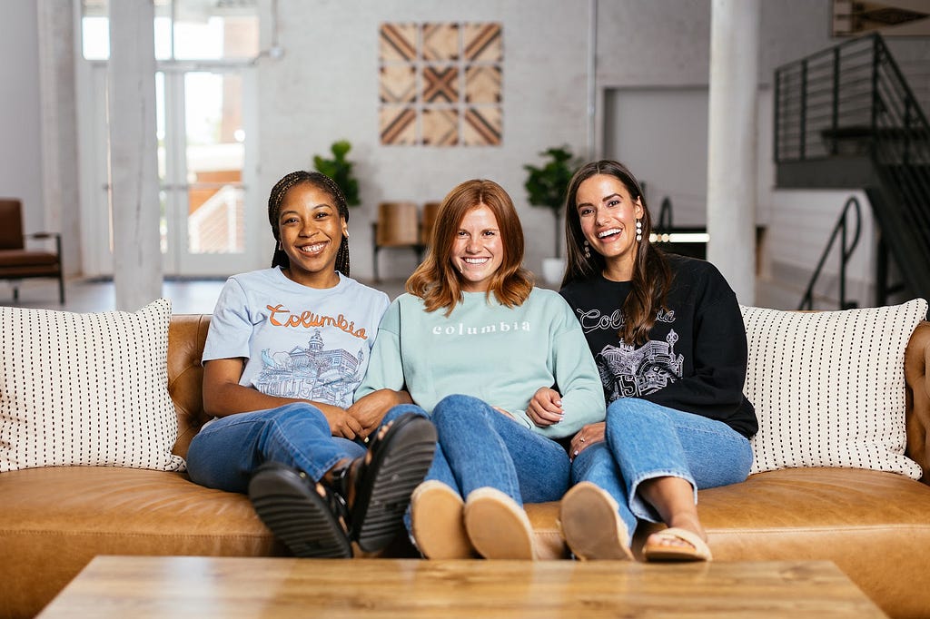 Three women, one Black and two white, sit on a leather couch in a bright, airy loft-style space. They model Six & Main long- and short-sleeved shirts with Columbia, South Carolina-specific designs on them, including the city name and landmarks.