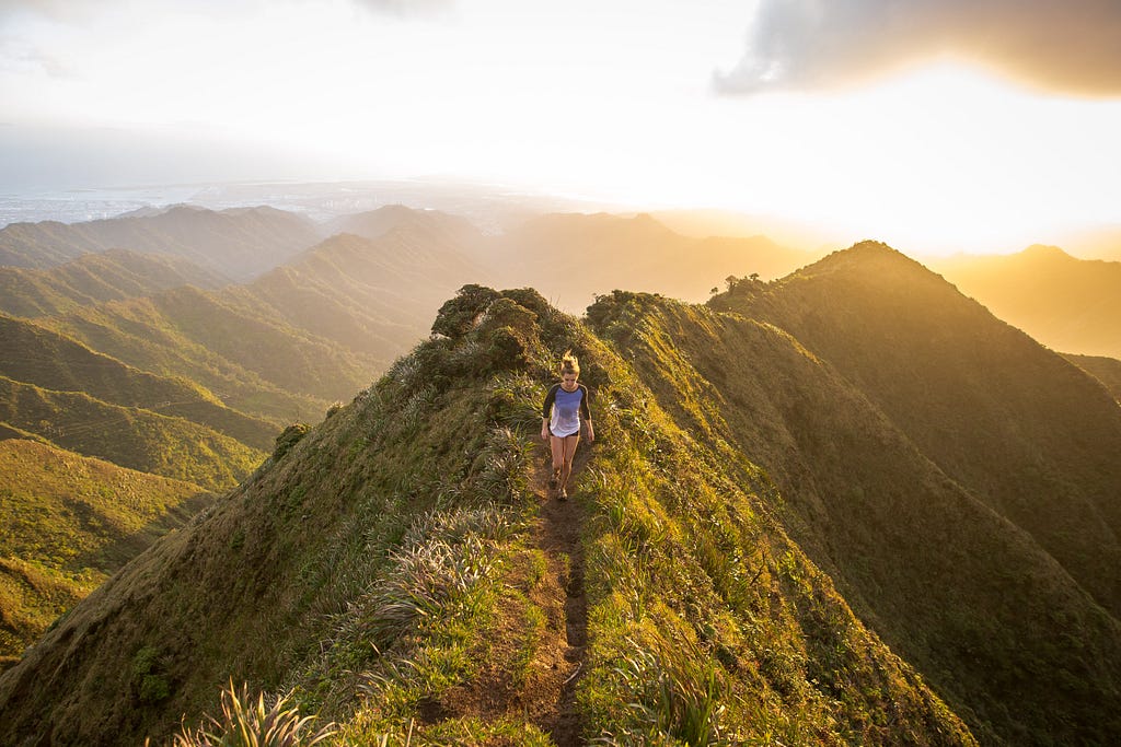 This image shows a girl doing trail running or hiking in the mountains over a green landscape with a beautiful sunset.