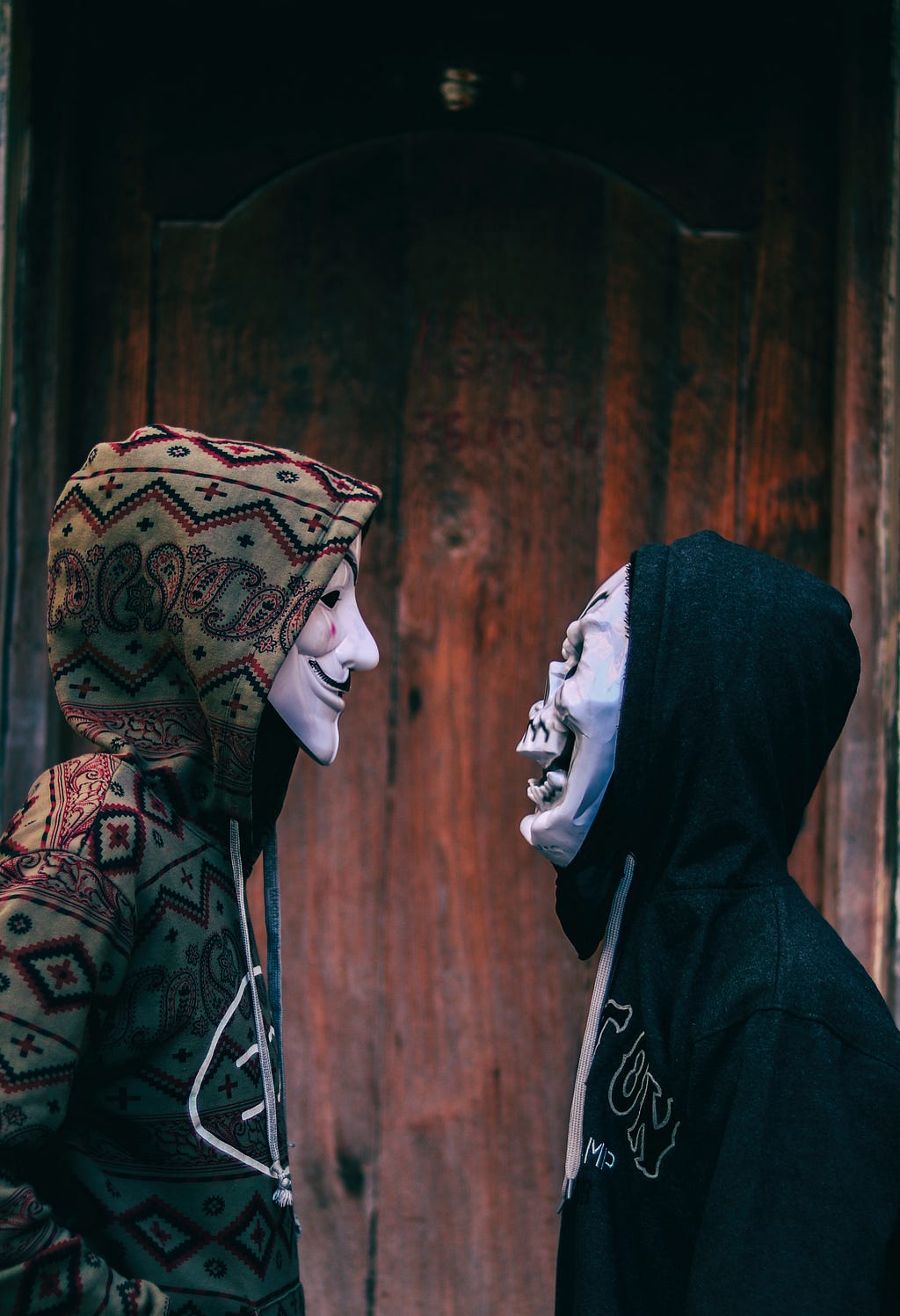 a man with a Guy Fawkes mask looking on a man with a skull mask