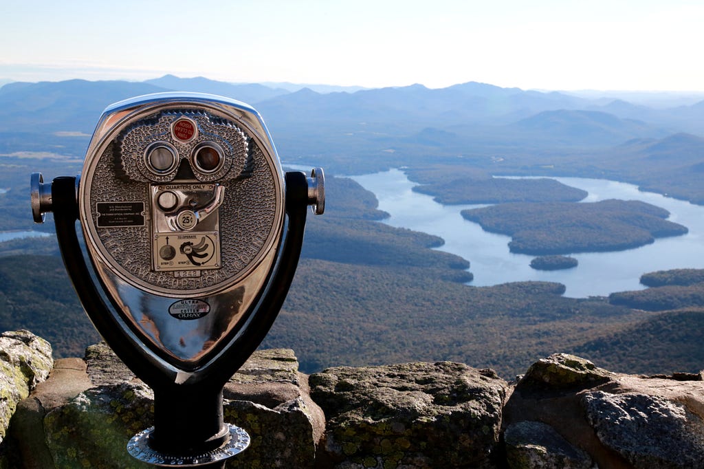 Par de binóculos posicionados à beira de um mirante alto. Ao fundo uma paisagem de um grande vale com florestas e rios.