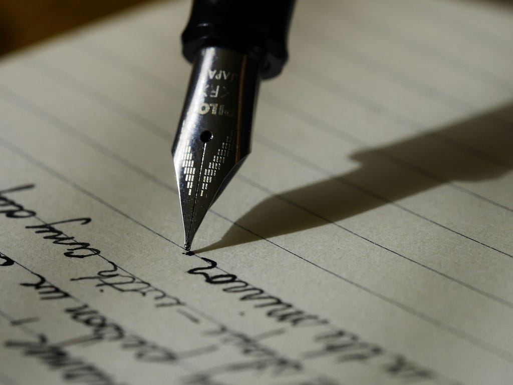 Picture of black cursive writing on lined paper, a fountain pen is being used.