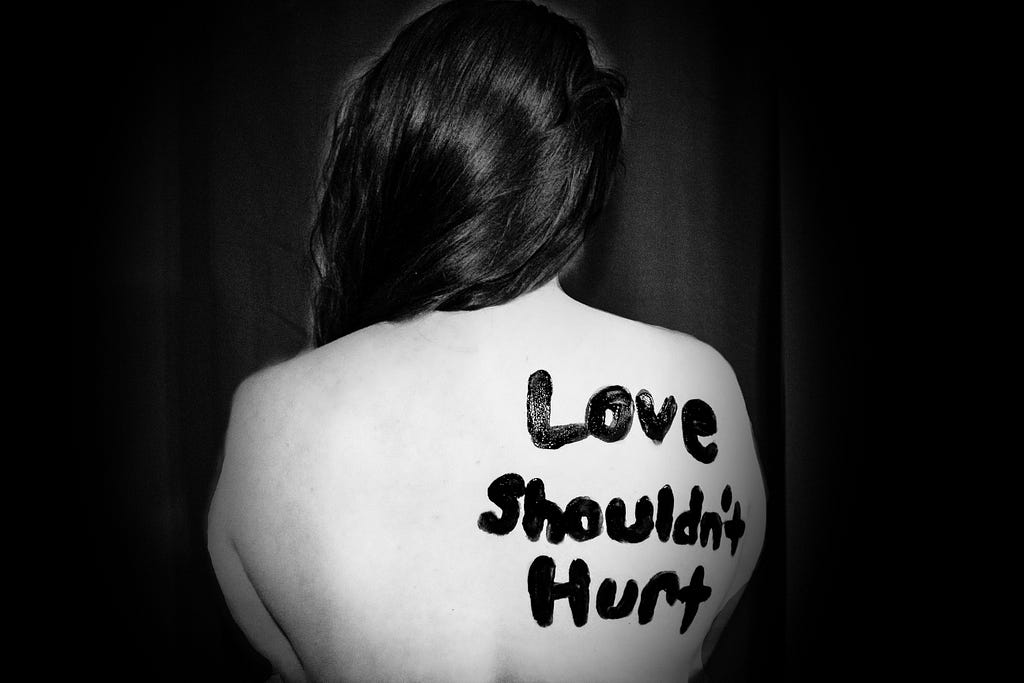 The back of a naked woman, with the words “Love shouldn’t hurt” painted on her skin.