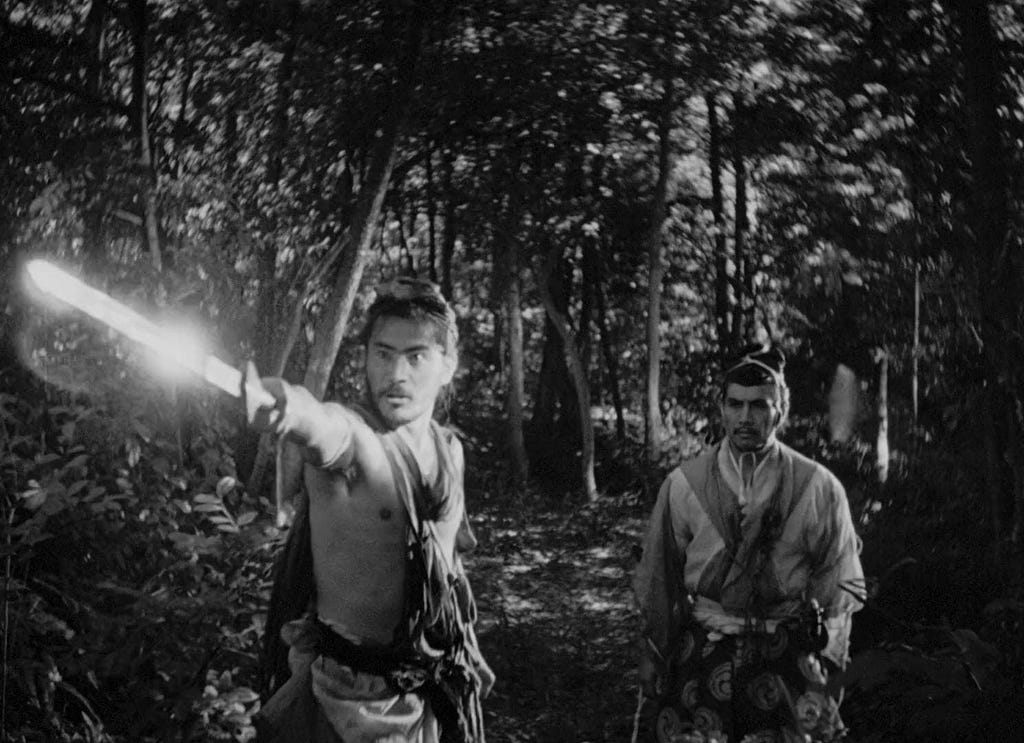 A random still from Rashomon (1950), showing the Bandit (Toshiro Mifune) and the Samurai (Masayuki Mori), in a forest. The Bandit has his sword drawn and is pointing it behind the camera. Both are dressed in the style of Medieval Japan, pre-Meiji restoration.