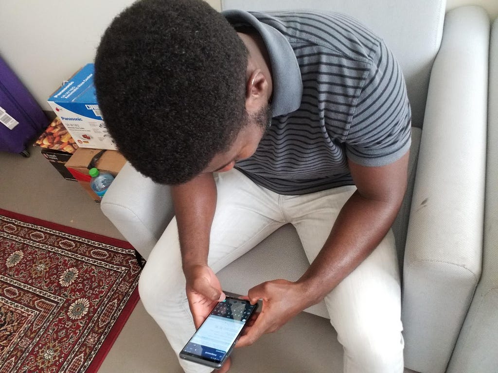 A young African man holds his smartphone. The screen shows a coding window. He leans over it and types with his thumbs.