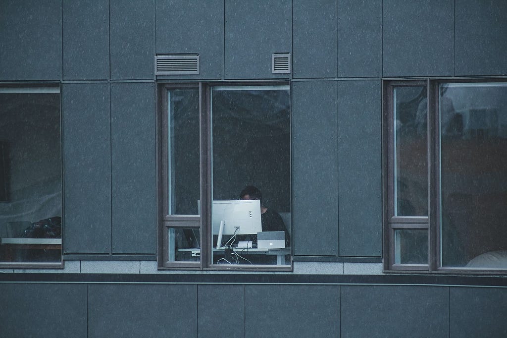 vista de fora de um homem trabalhando no computador em frente à janela. O prédio é cinza e o monitor se destaca por ser branco. | portainer ubuntu contêineres docker