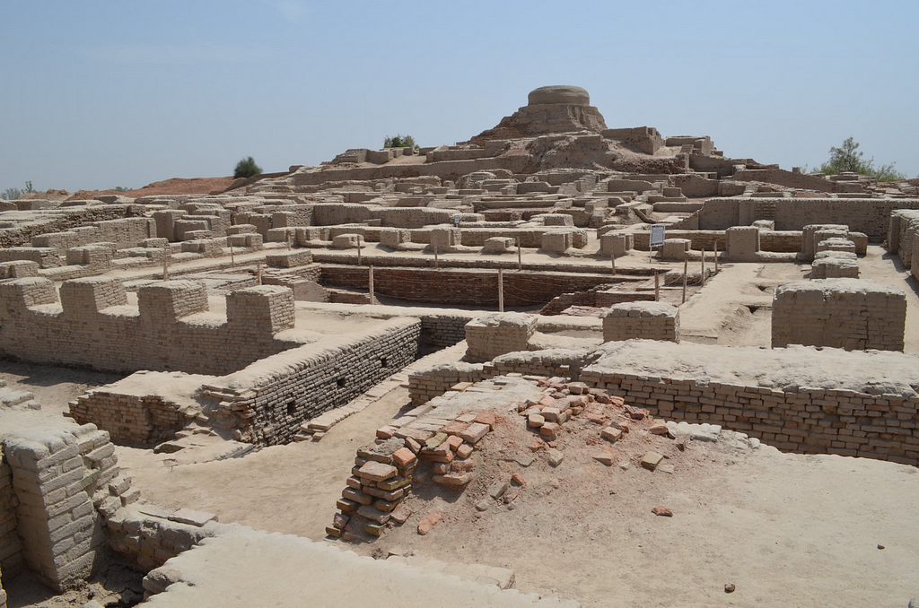 Ruins of Mohenjo-daro, an ancient Indus Valley Civilization city, featuring mud-brick structures and a large stupa-like structure in the background. The site showcases well-planned streets and buildings, reflecting advanced urban design from around 2500 BCE.