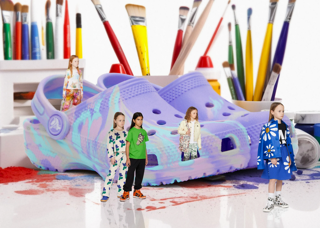 Kids wearing clothing from high fashion brand SSENSE are standing on a white table with paint behind massive purple Crocs shoes and colorful paint brushes in the background.