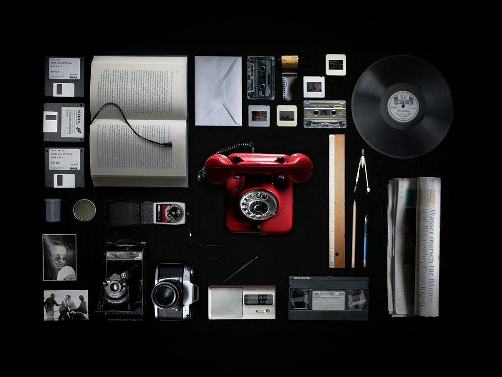 Old forms of media such as a rotary phone and film cameras displayed in a flat display on a black background