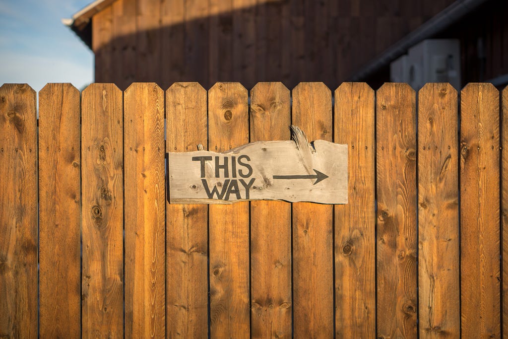 Wooden sign reading ‘this way’ with an arrow pointing to the right, mounted on a fence