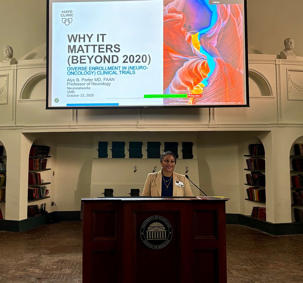 A woman stands behind a wooden podium delivering a presentation in a lecture hall. She is smiling and wearing a tan blazer over a dark top, with her hair styled neatly back into a bun. Above her, a large screen displays a slide titled &ldquo;Why It Matters (Beyond 2020): Diverse Enrollment in (Neuro-Oncology) Clinical Trials,&rdquo; with a colorful abstract medical image on the right. The room features arched walls and built-in bookshelves.