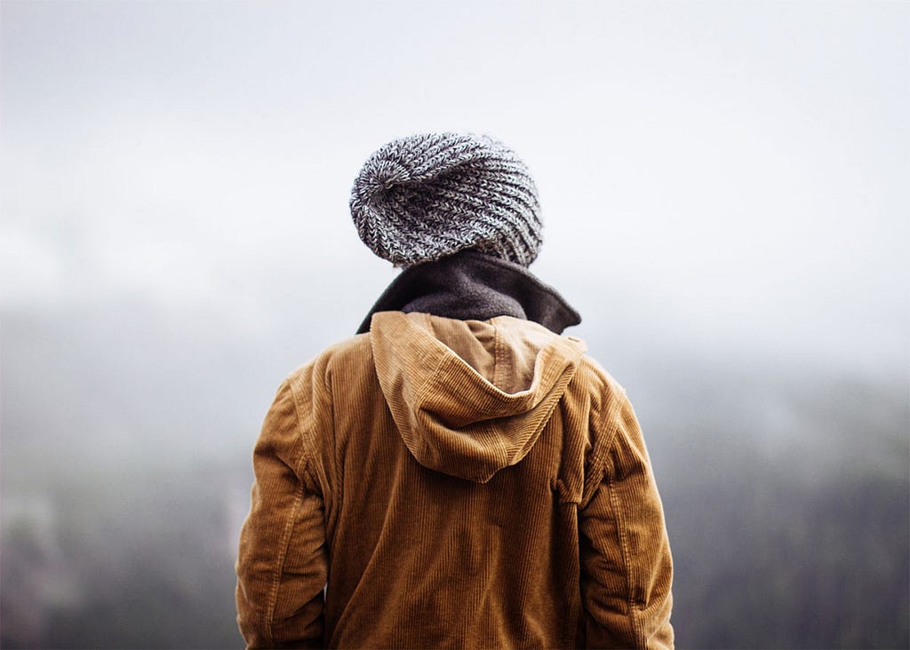 A person looking out over a foggy mountain landscape. We see them from the back and waist up, wearing a brown jacket and a grey knit cap.