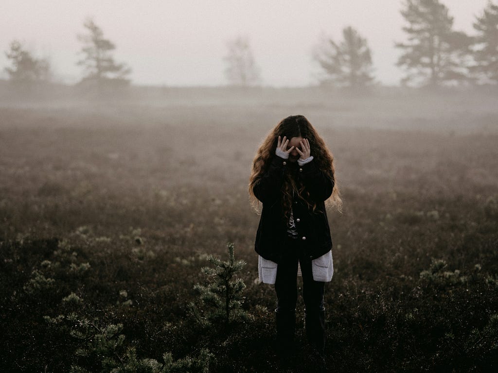 A woman, alone in a field, covers her face in an evident state of embarrassment