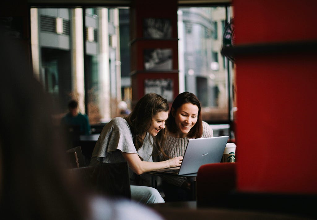 Two women in a cafe look at a laptop screen together.