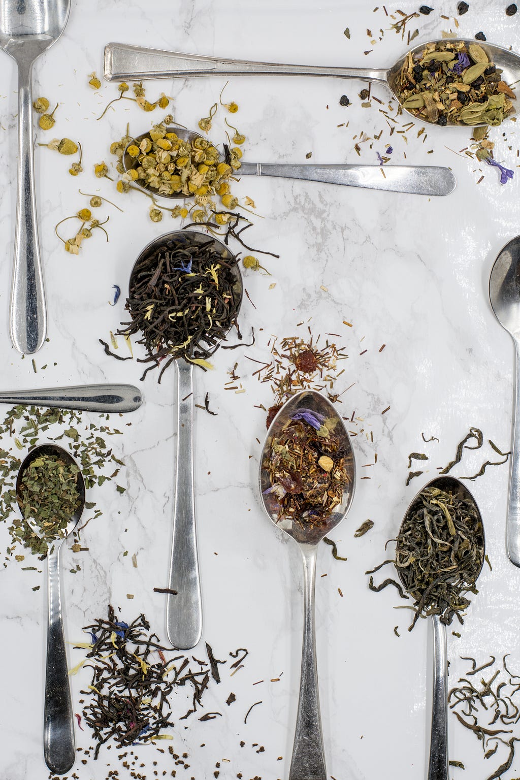 A selection of spoons laying on a white background, each spoon contains herbs or spices that are spilling over the edges