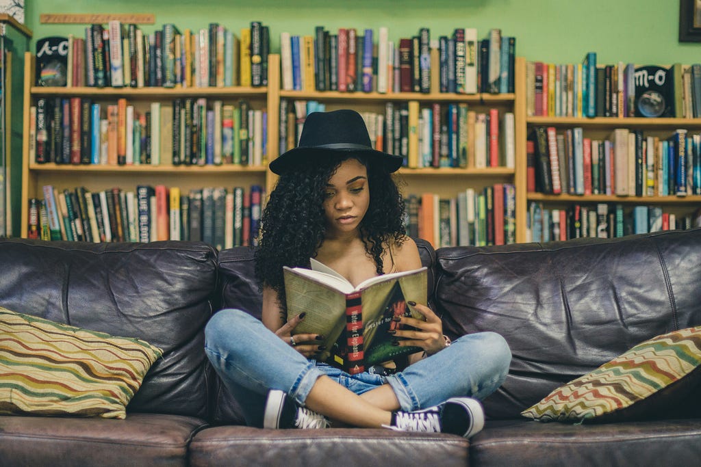 A girl sitting on a couch reading a book and a full shelf of books behind her.